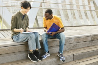 Two young adults study together outside on wooden steps. They are smiling and sharing notes. The