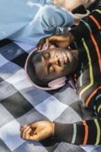Friends relaxes on a checkered blanket, wearing headphones and basking in the sunlight. The