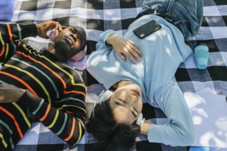 Two friends lie on a checkered blanket, wearing headphones, and enjoying music under the sun. A