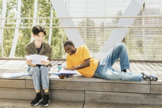 Two friends study outdoors on a sunny day, sitting and laying on a wooden steps surrounded by