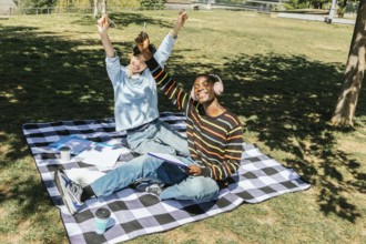 Two students friends are sitting on a checkered blanket in a sunny park. They are celebrating and