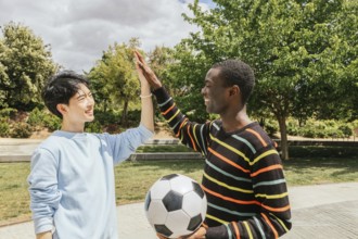 Two friends share a high five in a sunny park, celebrating after a soccer game. One holds a soccer
