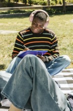 Friends sits on a checkered blanket in the sun, wearing headphones and a striped sweater, enjoying