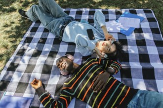 Two friends enjoy a sunny day outdoors, lying on a plaid blanket and listening to music with