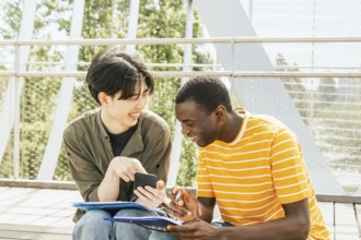 Two friends enjoy a sunny day outdoors while collaborating on a project. One holds a smartphone,