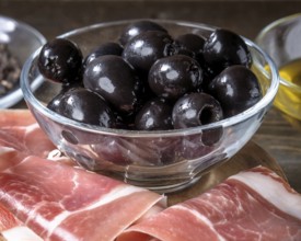 Juicy black olives in a glass bowl placed on a rustic wooden table, accompanied by thinly sliced