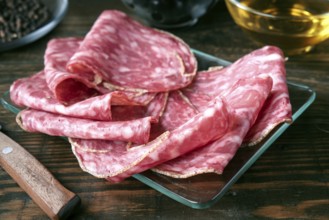Thin slices of spanish salami are arranged on a glass plate atop a rustic wooden table. The marbled