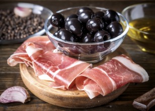 A rustic wooden board features thinly sliced spanish ham next to a glass bowl of black olives,