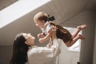 A joyful moment between a mother and her baby in a bright room. The mother lifts her smiling son in