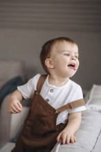 A cheerful toddler in brown overalls and a white shirt plays joyfully on a soft sofa. The playful
