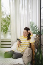 A woman in a striped yellow sweater relaxes on a bed using her smartphone. The room has a cozy
