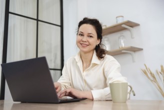 A professional woman, smiling and working on her laptop at a home office desk. Shelves and plants