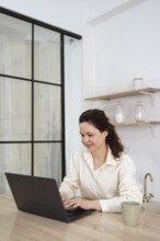A woman works from home, sitting at a table with a laptop. She is focused and looks at the screen,
