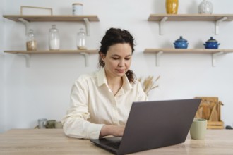 A woman sits at a table, focused on her laptop. Shelves with jars and decor create a cozy,
