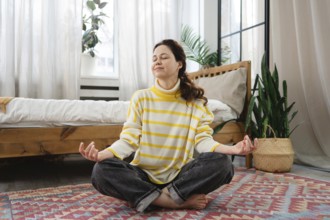 A woman sits cross legged on the floor of a cozy, sunlight filled bedroom at home, meditating in a