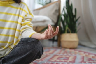 A woman meditates indoors, seated on a colorful rug. Their fingers are poised, surrounded by cozy,