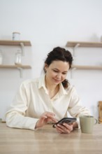 A woman in a cream shirt sits at a wooden table at home using her smartphone, next to a green