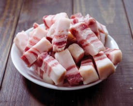A pile of salted pork belly cubes on a white plate displayed on a wooden table. Ideal for
