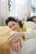 A woman in a striped sweater enjoys a relaxing moment at home. She lies on a bed in a sunlit room,