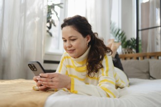 Smiling woman wearing a striped sweater lays on her bed and uses her smartphone. The bright and