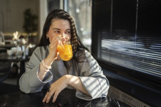 A mid-aged woman sips an orange drink in a brightly lit cafe, her expression serene and content as
