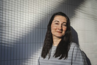 A middle-aged woman smiles gently while standing against a geometrically patterned tile wall, with