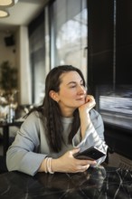 A middle-aged woman sits peacefully in a cafe, gazing outside while holding a smartphone in her