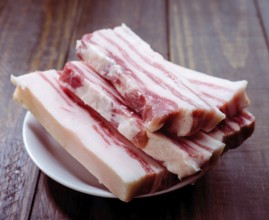 Slices of raw pork belly are neatly arranged on a white plate set against a rustic wooden surface.