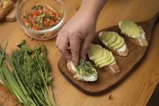 A hand garnishes avocado toast with fresh herbs on a wooden board. Nearby are a salad with tomatoes