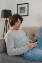 A young man with curly hair relaxes on a sofa, wearing a gray sweater, blue jeans, and headphones.