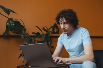 A young man with curly hair focused on his laptop in a modern room with orange walls and green