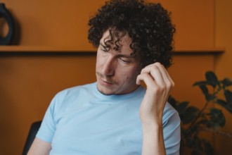 A young man with curly hair in a light blue shirt, sitting thoughtfully in a room with an orange