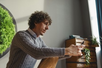 A young man with curly hair uses a smartphone, sitting beside a vibrant green wall. This scene