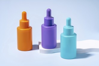 Three serum bottles in vibrant orange, purple, and turquoise colors, showcased against a soft blue