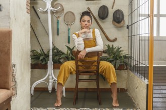 A young woman sits serenely on a wooden chair in a rustic home environment, surrounded by vintage