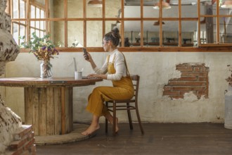 A young woman uses a smartphone and laptop while sitting at a round wooden table in a rustic cafe,