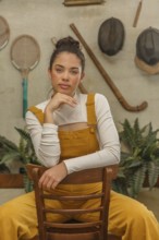 A young woman leans on a vintage wooden chair, dressed in a mustard yellow jumpsuit with a playful