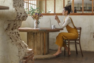 A woman uses a smartphone and laptop at a wooden table in a rustic setting, emphasizing the blend