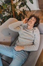 Young man with curly hair relaxes on a modern gray couch, wearing headphones and enjoying music. He