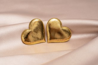 Two golden heart-shaped objects rest on a smooth pink satin surface, symbolizing love and