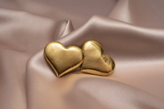 Two golden heart-shaped objects rest gently on a smooth, silky fabric, symbolizing love and