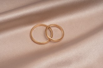 Two gold wedding bands gracefully overlap atop a blush satin surface, symbolizing eternal love and