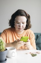 Woman at home, wearing facial beauty mask while browsing on her smartphone. A peaceful self-care