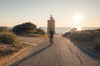 A man biking down a gravel road towards the historic Faro Camarinal lighthouse during sunset, with