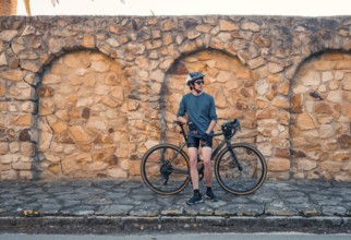 A young male cyclist stands next to his road bike, leaning against an aged stone wall in Zahara de