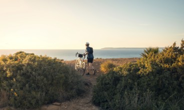 Back view of unrecognizable male cyclist standing with his bike, overlooking the ocean along a