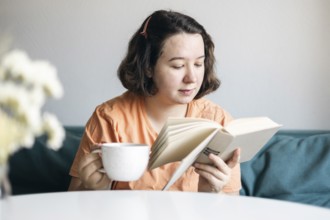 Young woman reads intently while holding a cup of coffee, comfortably seated on a sofa at home,