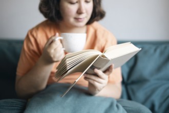 Cropped view of a woman sipping tea and reading a book, comfortably seated on a sofa