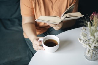 Cropped unrecognizable woman reading a book while holding a cup of coffee, seated comfortably at
