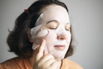 Image of a woman at home, using a facial pink quartz roller while wearing a sheet mask to enhance